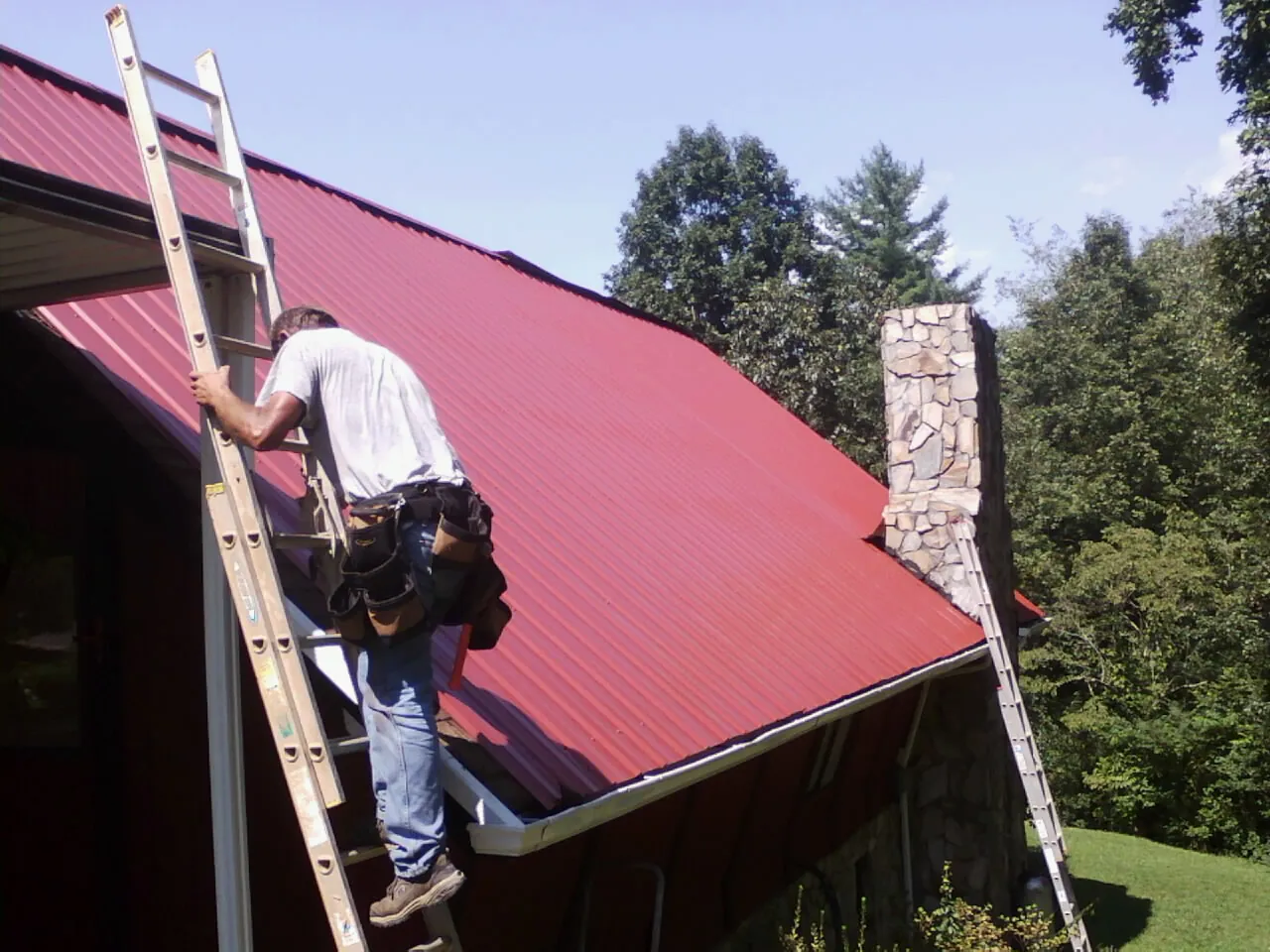 Roofer on a ladder beside a red metal roof and stone chimney — Soffit & Fascia Repair by Roofing Brothers in Troutdale